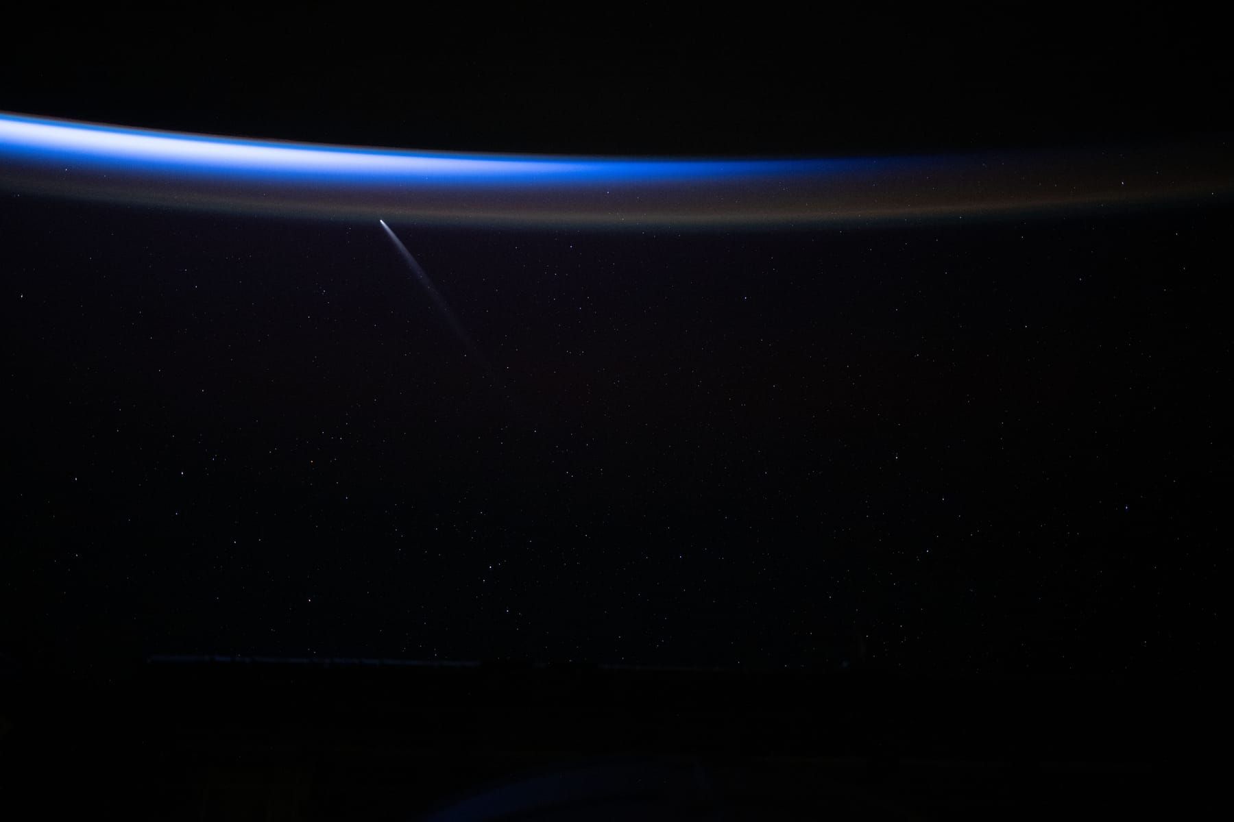 A comet with a straight tail above the blue band of Earth’s atmosphere, seen from orbit