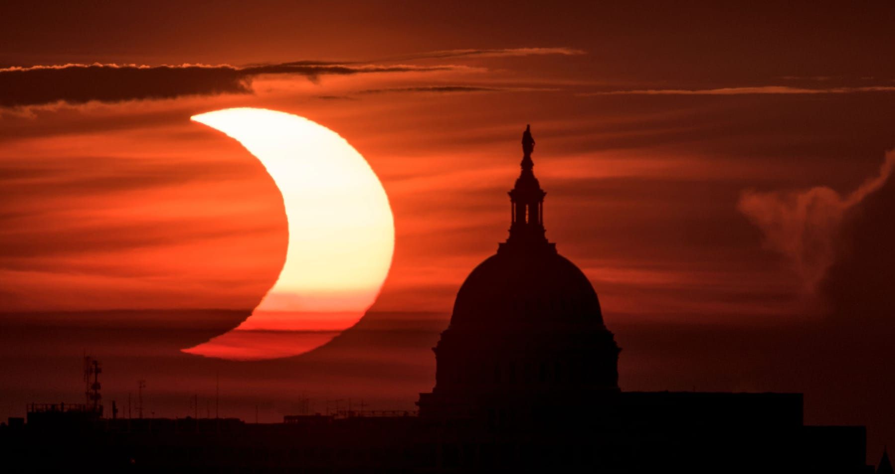 The crescent of a partially eclipsed Sun rising in an orange sky beside the dome of the US Capitol